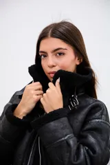 Campera tipo gamulán de corte oversize, confeccionada en cuero sintético con acabado texturizado y forro interior de corderito sintético negro. Presenta cierre frontal, cuello con solapas, detalles de tiras con hebilla en el ruedo y terminaciones en corderito en puños y bordes.