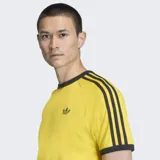 Remera de algodón Adidas Adicolor Sport de color amarillo, con cuello redondo y ribetes negros. Cuenta con las icónicas tres tiras negras en las mangas y el logo del trébol bordado en el pecho.