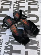 Sandalias de tacón alto Mutma modelo Ñoma, confeccionadas completamente en terciopelo color verde militar. Presentan una plataforma delantera, detalle de doble nudo o moña en la puntera, taco ancho y tira con hebilla ajustable en el tobillo.
