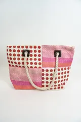 Bolso de playa o tote bag con diseño de patchwork en tonos rosados, naranjas y blancos. Presenta secciones con estampado de lunares rojos y secciones con tejido de rafia. Las asas son de cuerda trenzada.