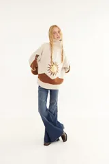 Sweater de punto en color beige con diseño de intarsia que representa un sol y montañas en tonos marrón y amarillo. Presenta cuello alto con cierre frontal, mangas largas y corte holgado.