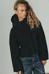 Cazadora acolchada de color negro con capucha, cuello subido y cierre frontal con cremallera y botones metálicos. Presenta manga larga con puño elástico, bolsillos delanteros y bajo ajustable con stoppers.