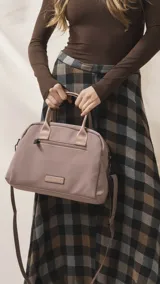 Cartera de mano color beige, confeccionada en tela impermeable, con doble asa corta y correa larga ajustable y desmontable. Presenta cierre principal, bolsillo frontal con cierre y detalle de colgante decorativo.