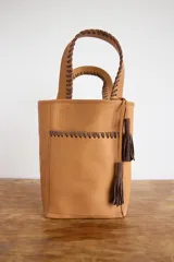 Matera tipo bolso de mano color tostado, con detalles de costuras y tiras de cuero marrón chocolate en las manijas y en el bolsillo exterior frontal. Incluye borlas colgantes decorativas.