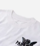 Remera blanca de algodón, corte oversized, con cuello redondo y mangas cortas. Presenta un estampado frontal con un ángel y el texto "PARADISE" en negro, y un pequeño gráfico de estrella/brújula en el lado izquierdo del pecho.