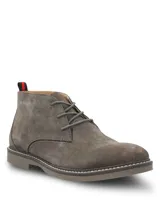 Botines tipo chukka Hush Puppies modelo New Safari, confeccionados en gamuza color gris. Presentan cierre de cordones, suela de goma con costura decorativa y detalle de tirador en el talón con franjas rojas y negras.