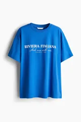 Remera de manga corta, cuello redondo y calce oversize, color azul vibrante, con estampa frontal en blanco que dice "RIVIERA ITALIANA Perla rara della costa LIGURIA, 1989".