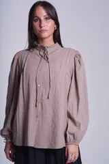 Camisa de color azul con cuello alto decorado con volados y lazo de ajuste. Presenta tablas decorativas en los hombros, cierre frontal con botones de nácar y mangas largas con puños elásticos que generan un efecto abullonado.
