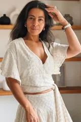Conjunto de blusa y pollera color beige con bordado artesanal. La blusa cuenta con escote en V cruzado y mangas cortas con volados, mientras que la pollera corta presenta diseño escalonado con volados.