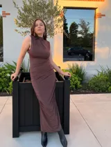 Vestido largo de color marrón, con diseño sin mangas y cuello alto. Presenta un detalle drapeado en el lateral que recorre la silueta, aportando textura y un ajuste entallado.