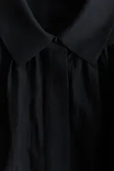 Blusa negra oversized en tejido liviano, con cuello clásico, tapeta con botones ocultos, canesú doble con frunces en el delantero y la espalda, y mangas globo largas con puños abotonados.