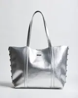 Cartera tipo tote de símil cuero plateado con doble asa de hombro, detalles de tachas en los laterales y placa metálica con logo de la marca en el frente.