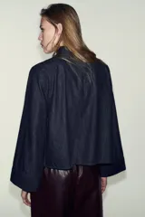 Camisa de denim azul oscuro, de corte cuadrado y manga ancha acampanada. Presenta cuello mao y cierre asimétrico con botones a presión en el frente.