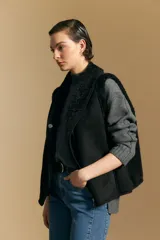 Chaleco negro de gamuza sintética con interior y bordes de corderito sintético. Presenta un diseño sin mangas con cuello solapa y cierre frontal.