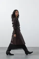 Trench coat de símil cuero color marrón oscuro, con solapas amplias, cierre frontal con botones, cinturón ajustable en la cintura y mangas con tiras ajustables. Presenta un largo por debajo de la rodilla y bolsillos laterales.