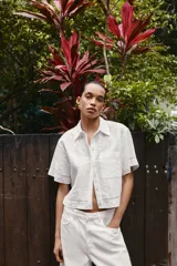 Camisa de algodón blanca con cuello solapa, manga corta y bolsillo de plastrón en el pecho. Presenta detalles de bordados a tono y cierre frontal con botones ocultos.