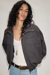 Chaqueta estilo bomber de color gris oscuro, con cuello subido, manga larga y corte holgado. Presenta bolsillos delanteros con solapa, bajo con pliegues y cierre frontal con cremallera oculta y botones a presión.
