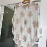 Camisa blanca de manga larga con estampado damasco en tonos camel, cuello camisero, cierre frontal con botones y diseño de corte asimétrico más largo en la espalda.