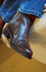 Botín negro de cuero liso, con puntera fina y taco bajo. Presenta costuras decorativas en hilo color crudo y cierre lateral con tirador.