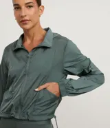 Campera cortavientos deportiva modelo cropped, color blanco traslúcido, con cierre frontal de cremallera, capucha estructurada, puños elásticos y bajo ajustable con cordón.
