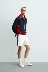 Chaqueta deportiva de corte regular, confeccionada en tejido piqué con interior forrado en red. Presenta un diseño color block en negro y rojo con detalles en blanco, cuello subido, cierre frontal con cremallera y bolsillos laterales con cierre.