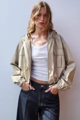 Cazadora color beige con capucha ajustable, manga larga con puño elástico, bolsillos frontales con solapa, bajo ajustable y cierre frontal con cremallera y botones a presión.