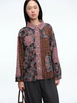 Camisa de manga larga con diseño patchwork que combina paneles de tela a cuadros en tonos rojizos, grises y amarillos con secciones de tela rosada estampada con flores pequeñas. Presenta cuello clásico, cierre frontal con botones y un bolsillo en el pecho.