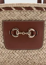 Bolso de mano y bandolera de fibra natural trenzada, color beige, con detalles en cuerina marrón y aplique metálico dorado en el frente. Cuenta con doble asa corta y correa larga desmontable.