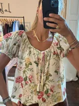 Camisa de lino color crudo con estampado floral en tonos rosados, amarillos y verdes. Presenta escote en V con cordones ajustables y mangas cortas abullonadas con elástico.