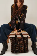 Bolso tote de algodón negro con estampado en tonos marrones y ocres, con costuras visibles en color claro. Presenta tiras de hombro negras y un adorno colgante con borlas negras y detalles dorados.