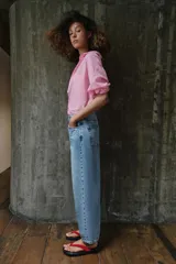 Pantalón de jean tiro medio con diseño barrel, caracterizado por su pernera holgada que se estrecha hacia el tobillo. Presenta cinco bolsillos, cierre frontal con cremallera y botón, y detalle de raya planchada en el frente.