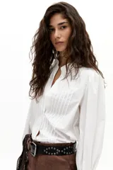 Camisa blanca de popelina de algodón con alforzas en el frente, cuello con terminación ondulada y bordados, y puños con vainillas. Tiene un corte levemente entallado, ruedo curvo y un sutil frunce en los hombros.