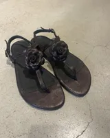 Sandalia chata de cuero color marrón oscuro con acabado metalizado. Presenta un diseño de tiras tipo thong con una flor tridimensional aplicada sobre la tira central. Posee tira fina ajustable con hebilla en el tobillo.