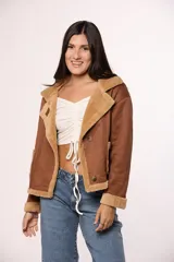 Campera tipo gamulán de gamuza color tostado con interior y solapas de corderito sintético color crema. Presenta cierre frontal con botones, bolsillos cuadrados delanteros y detalles de costuras visibles.