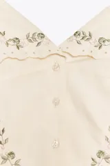 Top de color crema con escote en pico, tirantes anchos y cierre frontal de botones. Presenta bordados florales en tono verde claro alrededor del escote y en forma de semicírculo en la parte inferior delantera.