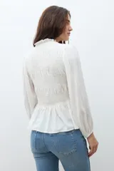 Camisa de color crudo con detalle de fruncido en el pecho, cuello redondo con volado sutil, cierre frontal con botones y mangas largas abullonadas con puños elásticos.