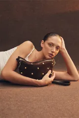 Bandolera de gamuza color marrón con diseño de media luna, decorada con tachas metálicas doradas distribuidas en el frente. Cuenta con cierre superior metálico y correa larga ajustable para llevar cruzada.