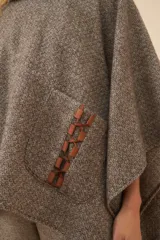 Poncho de lana color beige con cuello alto, bolsillos frontales y detalle de cordones de cuero en los bordes de los bolsillos.