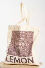 Bolsa tipo tote bag color crudo, con tiras de hombro, confeccionada en 100% algodón. Presenta un diseño gráfico frontal con estampado en tonos morados que incluye texto con las palabras "fitness, wellness, gym" y la marca "LEMON" en la parte inferior.