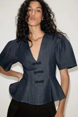 Blusa de denim oscuro con escote en pico, mangas cortas abullonadas y diseño peplum con bajo evasé. Presenta cierre frontal con alamares decorativos y cierre lateral con cremallera oculta.