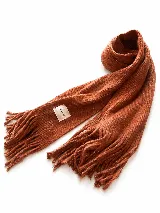 Bufanda tejida de textura suave y cálida, color terracota, con flecos en los extremos y etiqueta decorativa con la inscripción 'HAND MADE'.