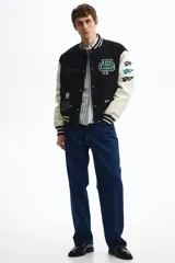 Campera estilo varsity con cuerpo negro y mangas en contraste color crema. Presenta múltiples parches y bordados decorativos en el frente y las mangas, cuello, puños y ruedo acanalados a rayas, y cierre frontal con botones a presión.