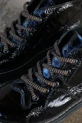 Borcegos de cuero charolado negro con cordones jaspeados en tonos marrones y suela track dentada con costura blanca visible.