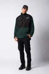 Campera de polar con recortes en tela técnica tipo sire en la parte superior frontal. Presenta cierre frontal completo, cuello alto y bolsillos funcionales en el pecho. Diseño bicolor en tonos verde oliva y negro con logo de la marca en el pecho.