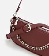 Cartera tipo baguette color bordó, confeccionada en poliuretano con acabado liso. Presenta una cadena decorativa plateada en el frente, cierre superior con cremallera y correa larga ajustable y desmontable.