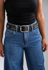 Cinto negro de cuero sintético con textura graneada, hebilla metálica rectangular plateada y apliques de tachas metálicas en los laterales.