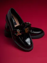 Mocasín de charol negro con plataforma, detalle de lazo y aplique de oso dorado.