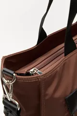 Bolso tipo tote color negro, con doble asa superior y correa desmontable. Presenta un bolsillo frontal con solapa, bolsillos laterales y un detalle colgante decorativo con tiras de colores.