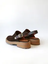 Zueco de cuero gamuzado color chocolate, con diseño de punta cerrada y tira ajustable con hebilla metálica en el talón. Presenta una suela de plataforma dentada en tono marrón claro.