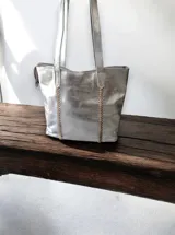 Cartera plateada tipo tote bag de cuero sintético con doble asa de hombro y detalle de costuras laterales.